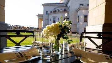 A table set for dining with a vase of flowers, clear glasses, and plates, overlooking a building.