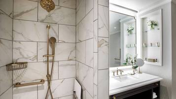 A bathroom with a marble tiled shower, gold fixtures, a white sink, and a mirror with a shelf of toiletries.