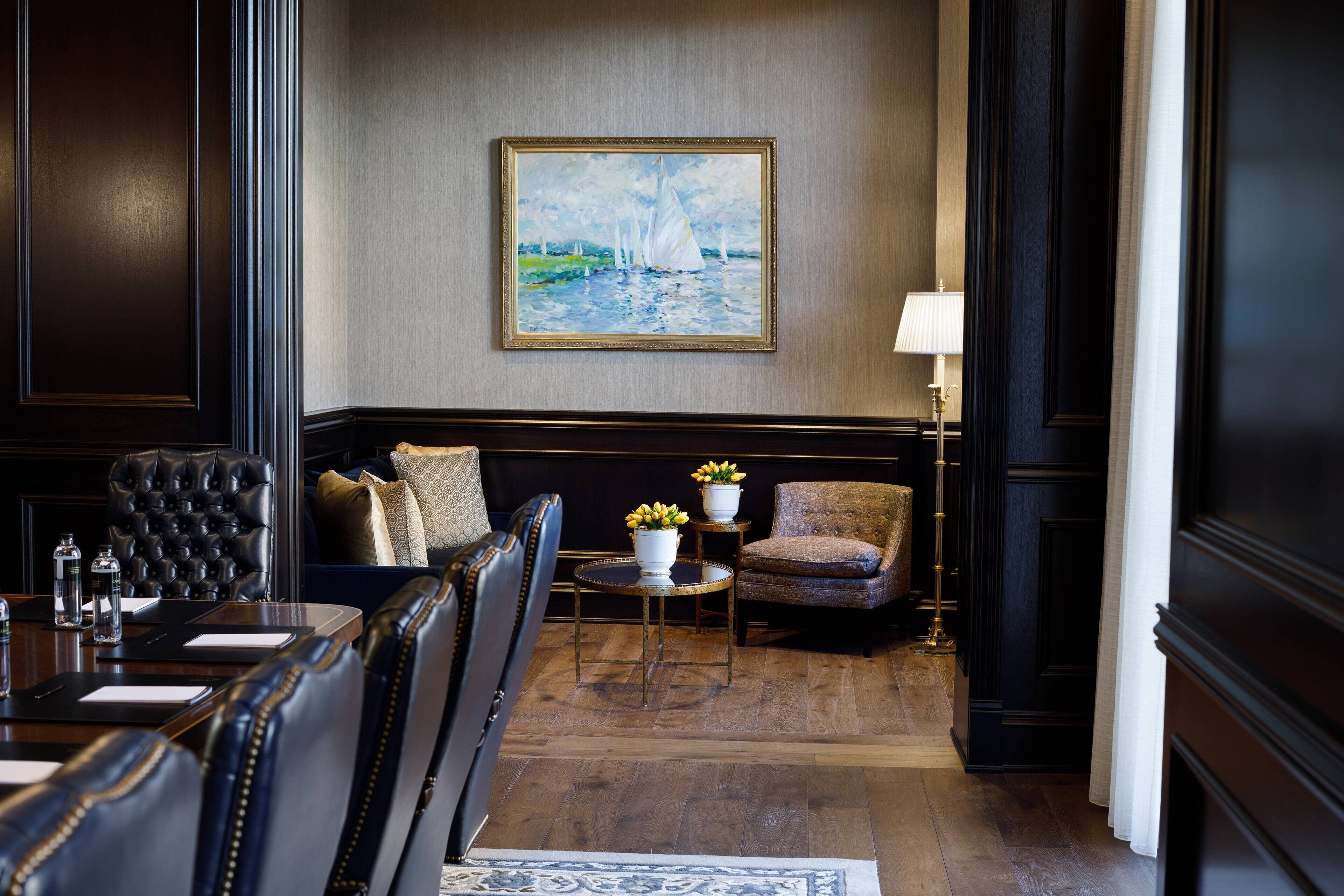 A hotel room with a dark wood paneling, a painting of a sailboat on the wall, a round table with a vase of flowers, and a sofa with cushions.