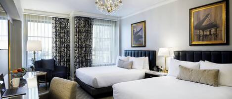 A hotel room with two beds, a desk with a laptop, a chair, a chandelier, and artwork on the walls.