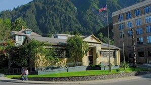 A building with a stone facade, a flagpole with an American flag, and a mountain backdrop. - Nature Retreat in Juneau-Near Town, Sleeps 4. (Juneau)