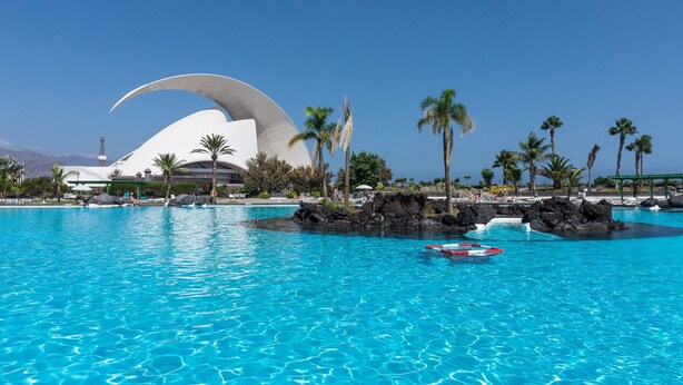 A large swimming pool with clear blue water, surrounded by palm trees and a distinctive white building with a curved roof. - HomeForGuest Design Attic Apartment, FREE parking (Santa Cruz de Tenerife)