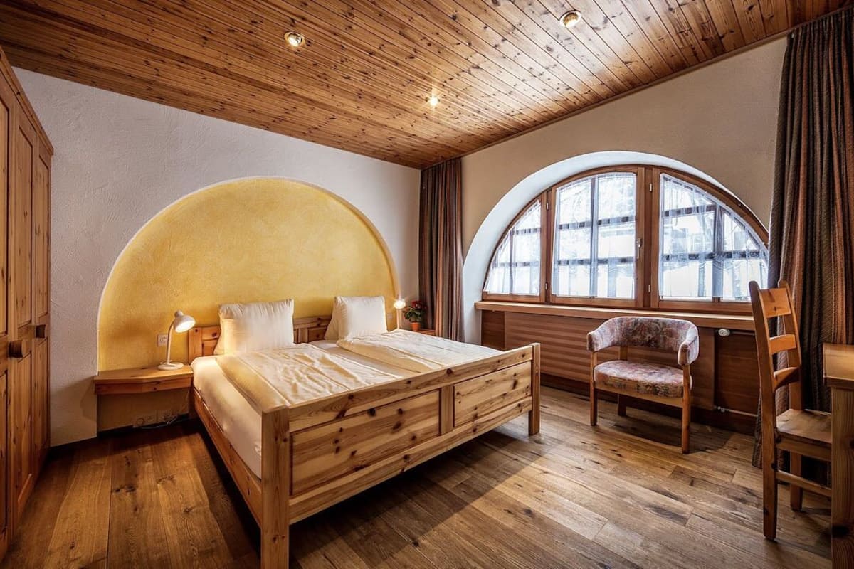 Une chambre avec un cadre en bois, un grand lit, une fenêtre cintrée et une chaise en bois.