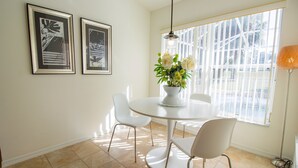 Un coin repas avec une table ronde, des chaises blanches et un vase rempli de fleurs. Deux tableaux encadrés ornent le mur, et une fenêtre est équipée de stores.