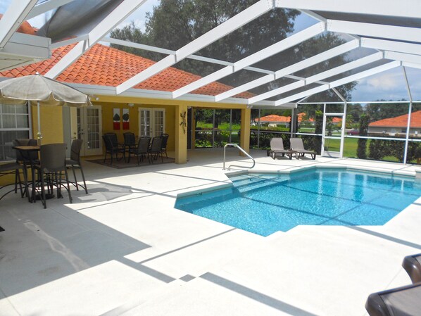 A covered outdoor pool area with a table and chairs, a pool ladder, and a view of a red-tiled roof building in the background. - Stylish Pool Close To Withlacoochee Bike Trail 3 Bedroom Home by RedAwning (Inverness)