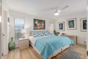 Un dormitorio con cama, mesitas de noche, ventilador de techo, ventana con persianas, un florero e impresiones botánicas enmarcadas en la pared.