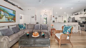 A spacious living room with a grey sofa, a coffee table, and a wicker chair. A kitchen with white cabinets and a stainless steel oven is visible in the background.