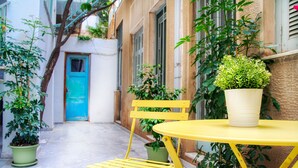 A narrow courtyard with a yellow folding chair and a small round table, surrounded by potted plants and a blue door. - Spacious 3-Bedroom Apartment Near the Train (Athens)