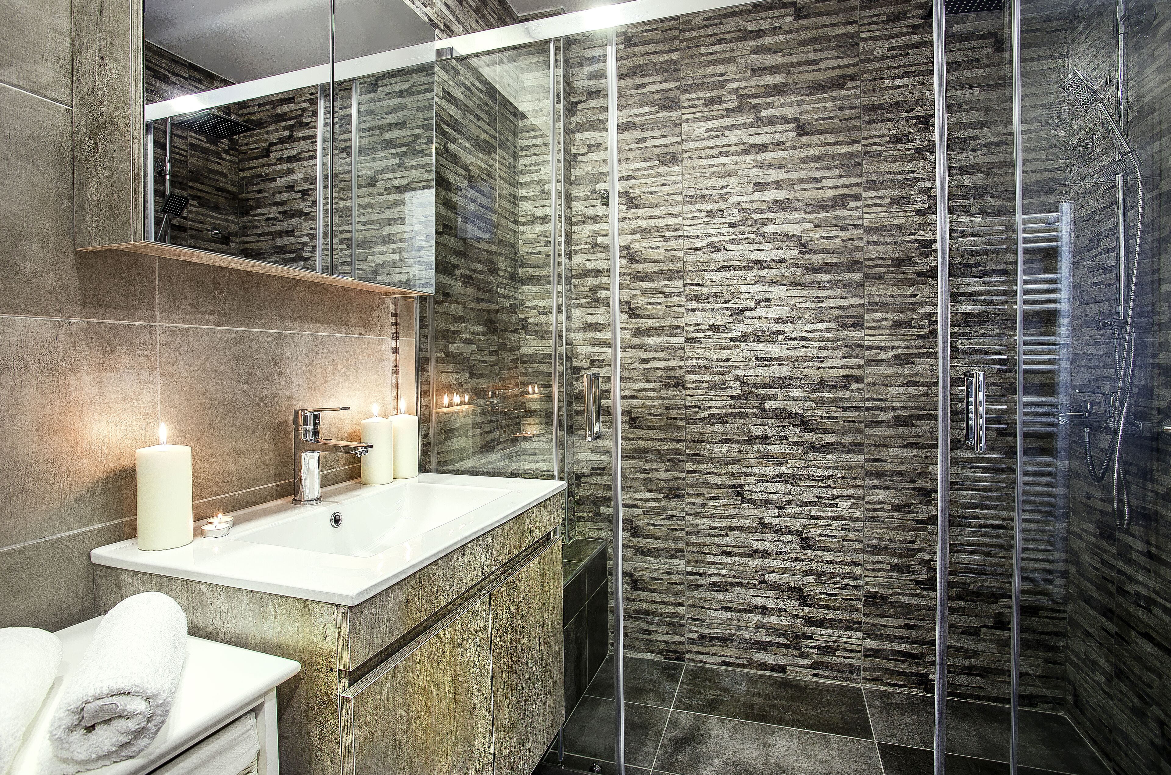 A modern bathroom with a stone-textured wall, a glass-enclosed shower, and a vanity with a mirror and candles.