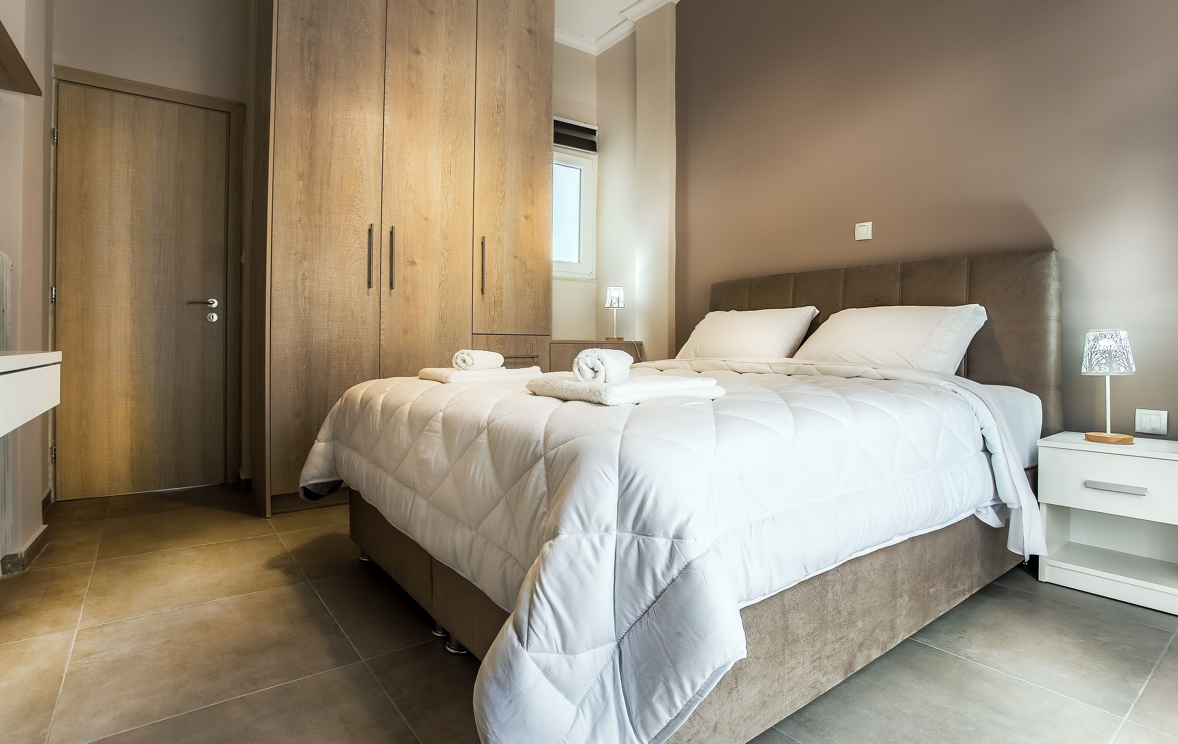 A bedroom with a large bed, a wooden wardrobe, a bedside table with a lamp, and a bathroom door.