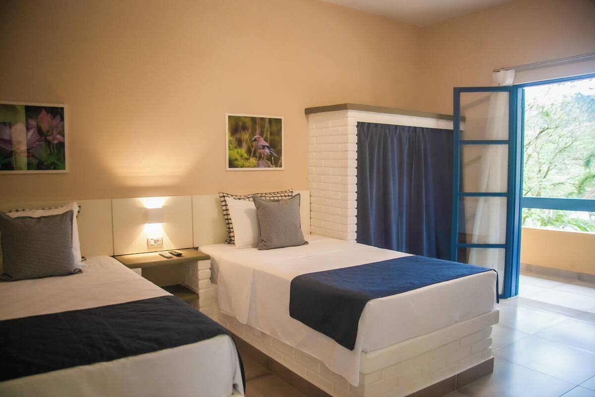 Una habitación de hotel con dos camas, una mesita de noche con lámpara, una ventana con cortinas y un cuadro de un pájaro en la pared.