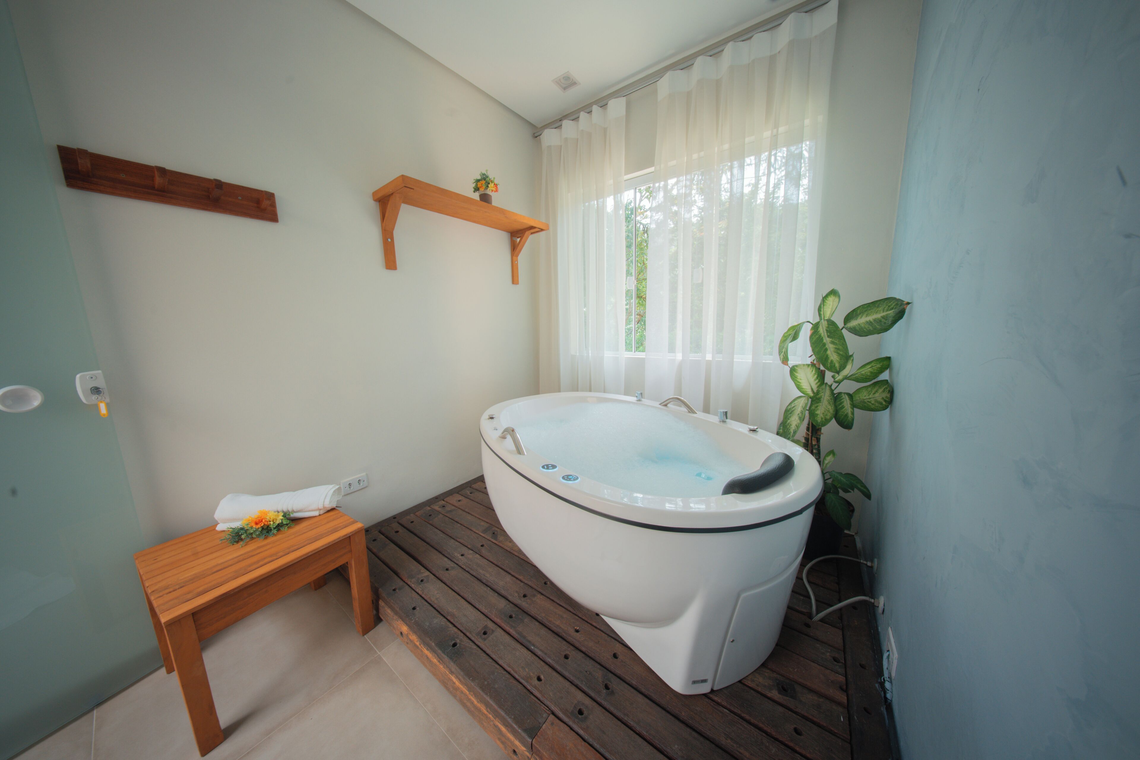 A bathroom with a wooden bench, a white bathtub, a wooden shelf with a plant, and a window with sheer curtains.