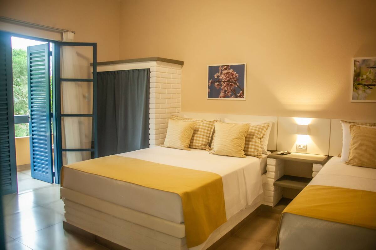 Una habitación de hotel con una cama grande, una mesita de noche con lámpara, una ventana con cortinas y un cuadro en la pared.