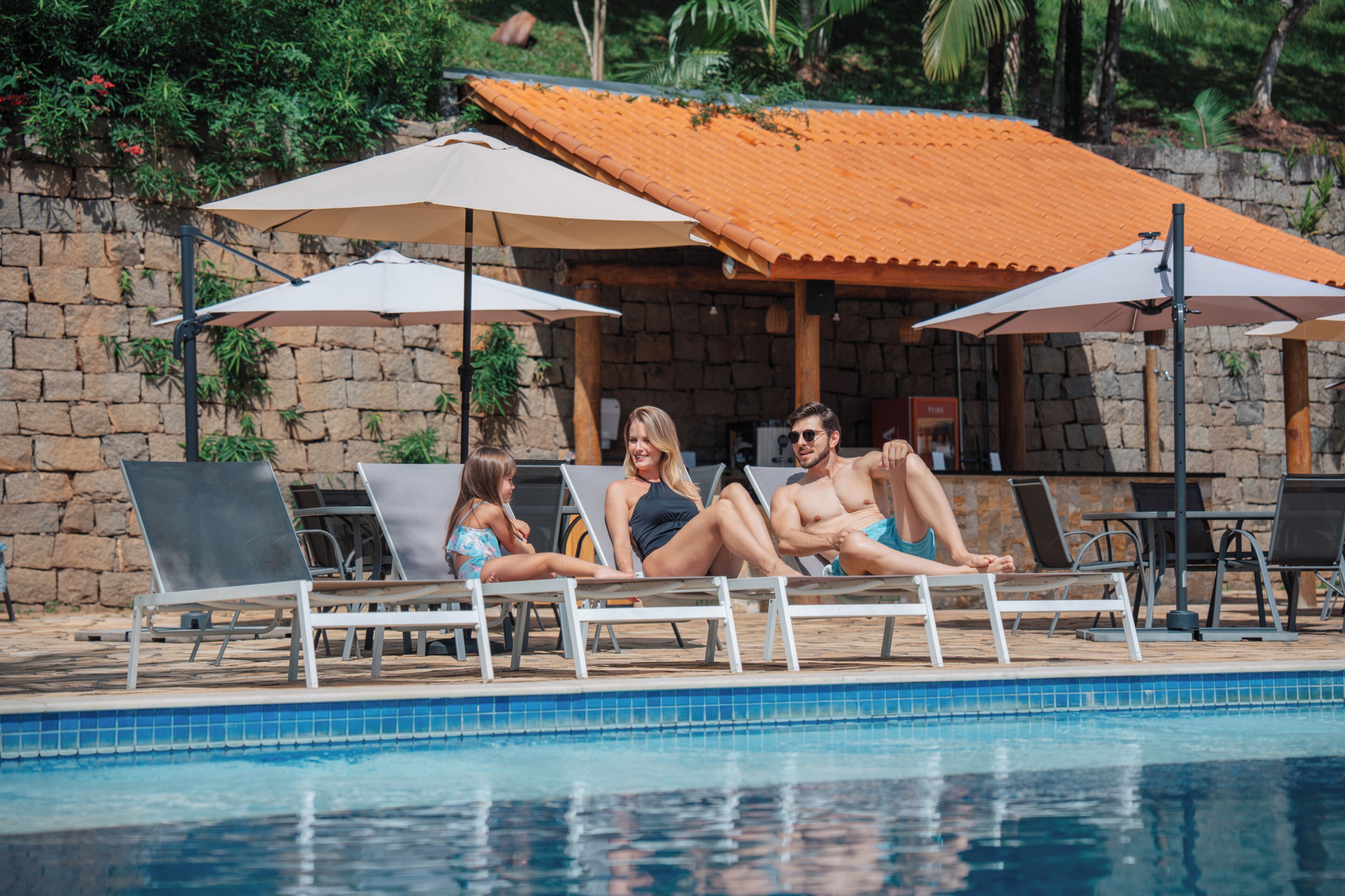 Un área junto a la piscina con sillones de descanso y sombrillas, una pared de piedra y una piscina con baldosas.