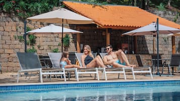 Un área junto a la piscina con sillones de descanso y sombrillas, una pared de piedra y una piscina con baldosas.