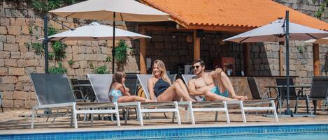 Un área junto a la piscina con sillones de descanso y sombrillas, una pared de piedra y una piscina con baldosas.