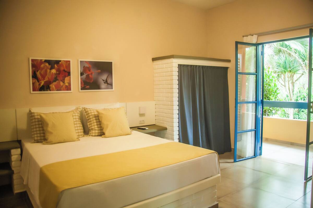 Habitación de hotel con cama, mesita de noche, ventana con cortinas y vista a la vegetación.