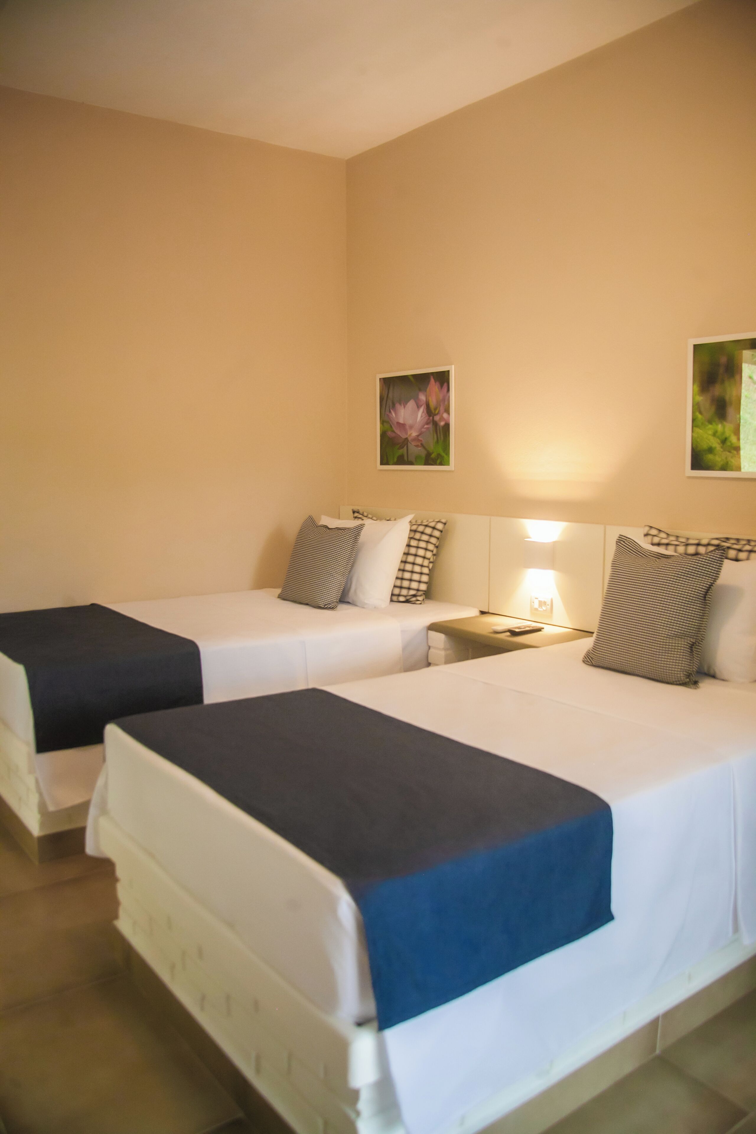 Habitación de hotel con dos camas individuales, cada una con una almohada y una manta. Hay una mesita de noche con una lámpara entre las camas. Las paredes son color beige, y hay dos cuadros enmarcados en la pared.