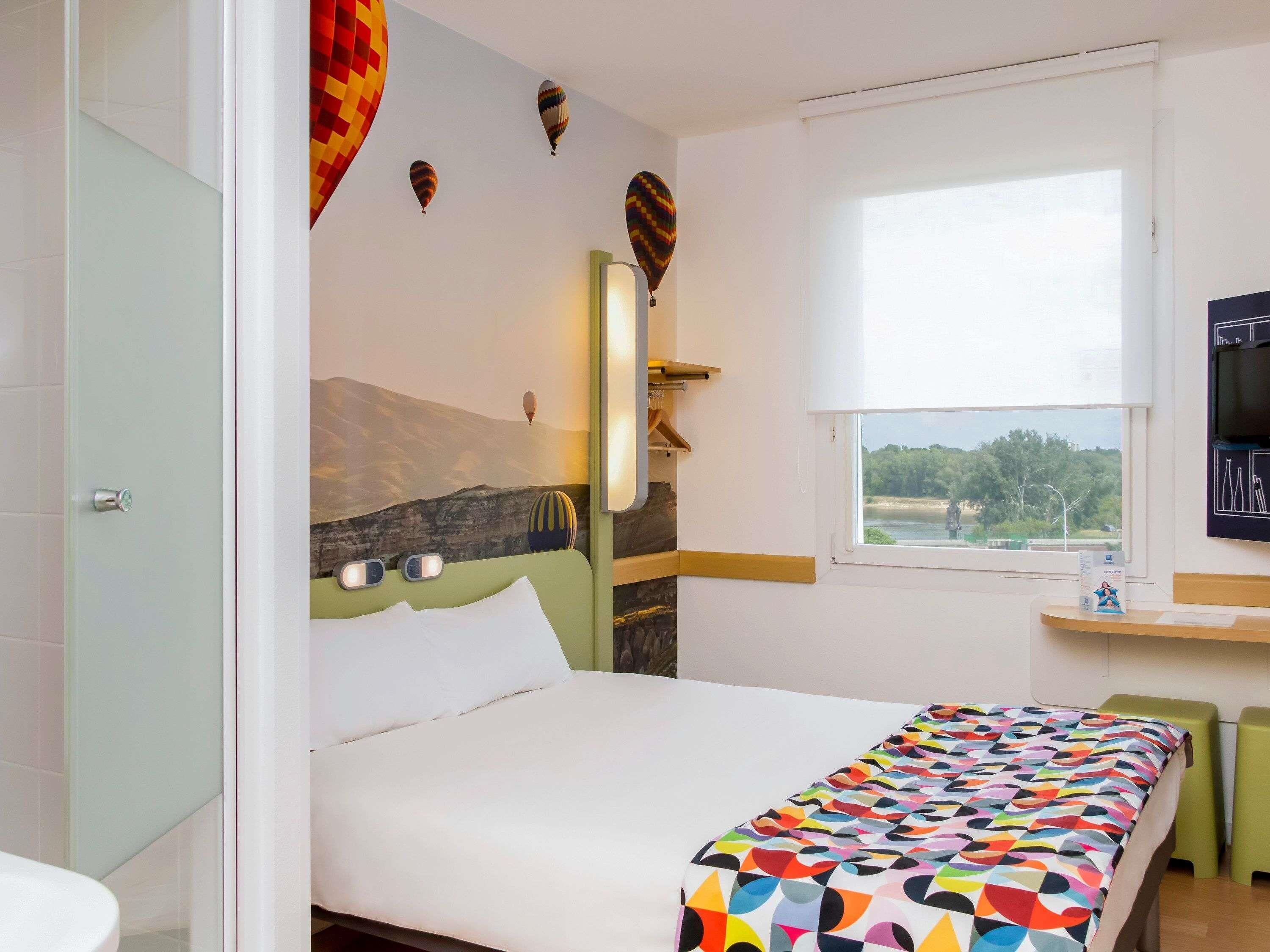 Habitación de hotel con cama, ventana con vistas, televisor y un cuadro de paisaje montañoso en la pared.