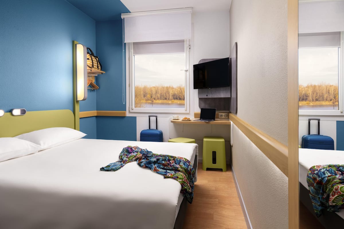 A compact hotel room with two beds, a small kitchenette, a TV, and a window with a view of trees.