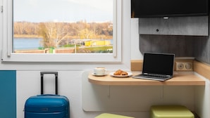 Habitación de hotel con escritorio, laptop, televisión, maleta y ventana con vista a un lago y árboles.
