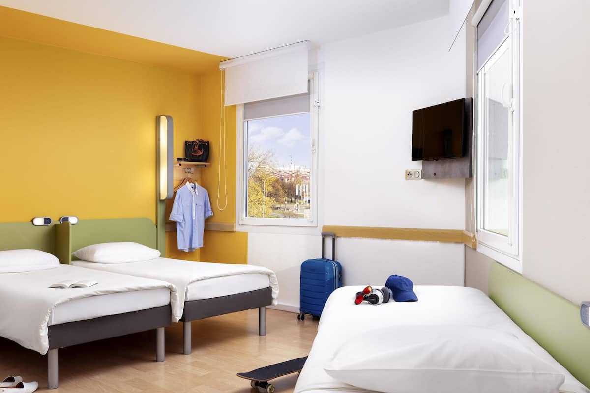 A hotel room with two beds, a window with a view, a TV mounted on the wall, and a suitcase on the floor.
