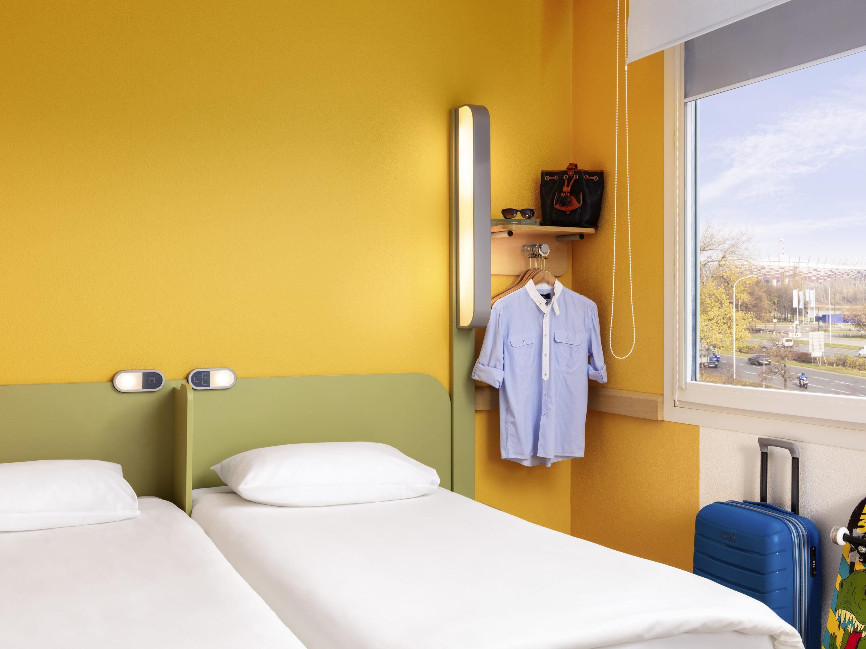 Habitación de hotel con dos camas, una ventana que ofrece vistas de la ciudad, un abrigo colgado y una maleta.