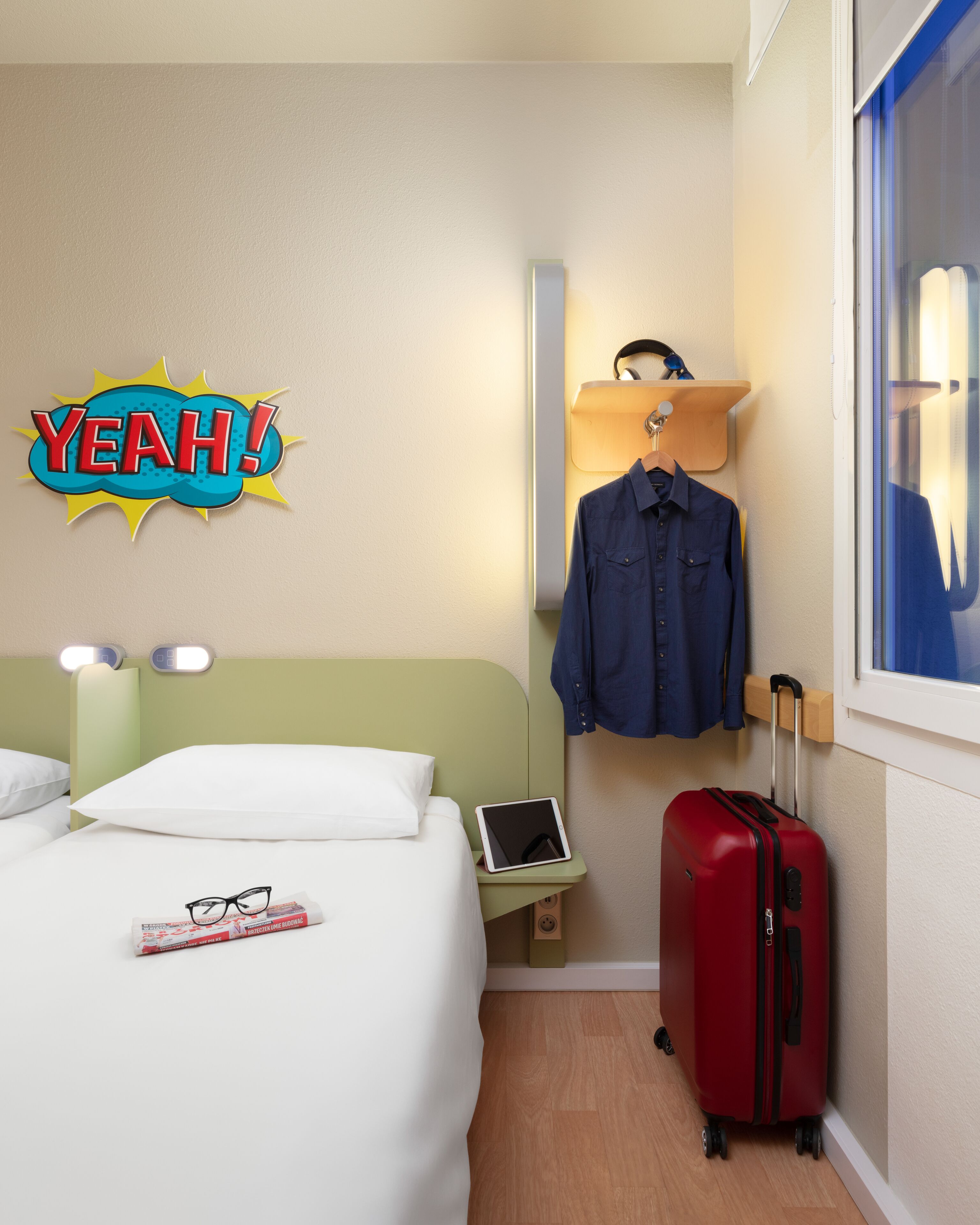 A hotel room with a bed, a bedside table with a tablet, a red suitcase, a blue shirt on a hanger, and a 'YEAH!' sign on the wall.