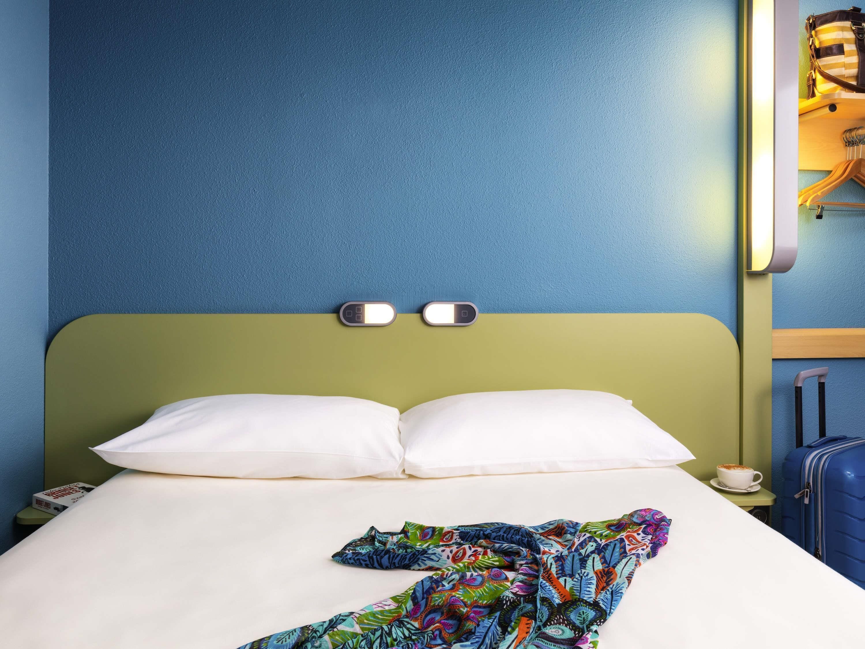 A hotel room with a bed, a nightstand, a lamp, a suitcase, and a cup of coffee.