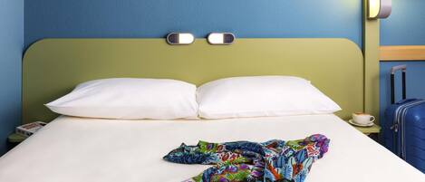 A hotel room with a bed, a nightstand, a lamp, a suitcase, and a cup of coffee.
