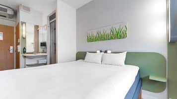 A hotel room with a large bed, a green headboard, a wall-mounted TV, and a bathroom visible through an open door.