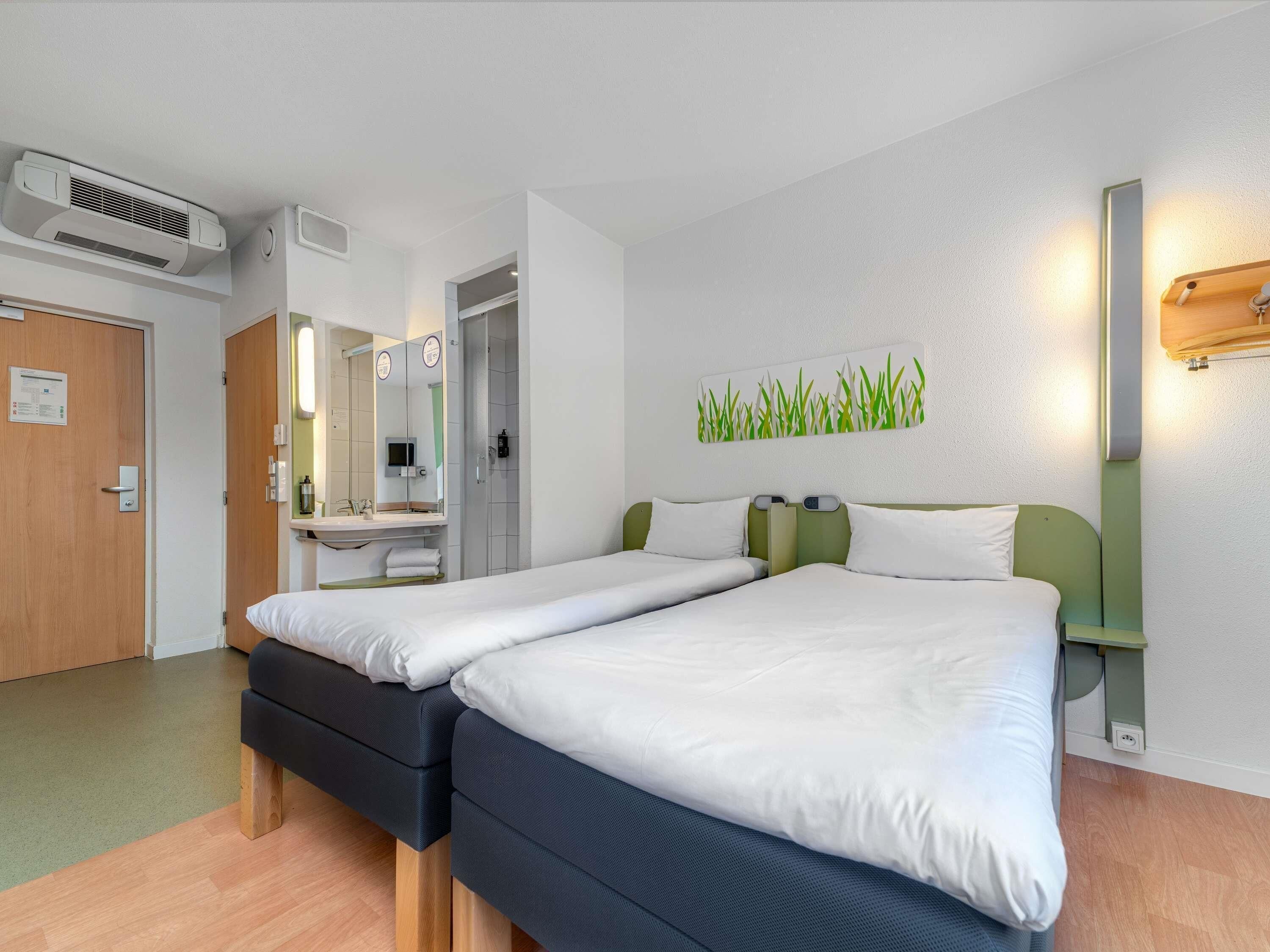 Une chambre d’hôtel avec deux lits, une salle de bain avec un miroir et un luminaire fixé au mur.
