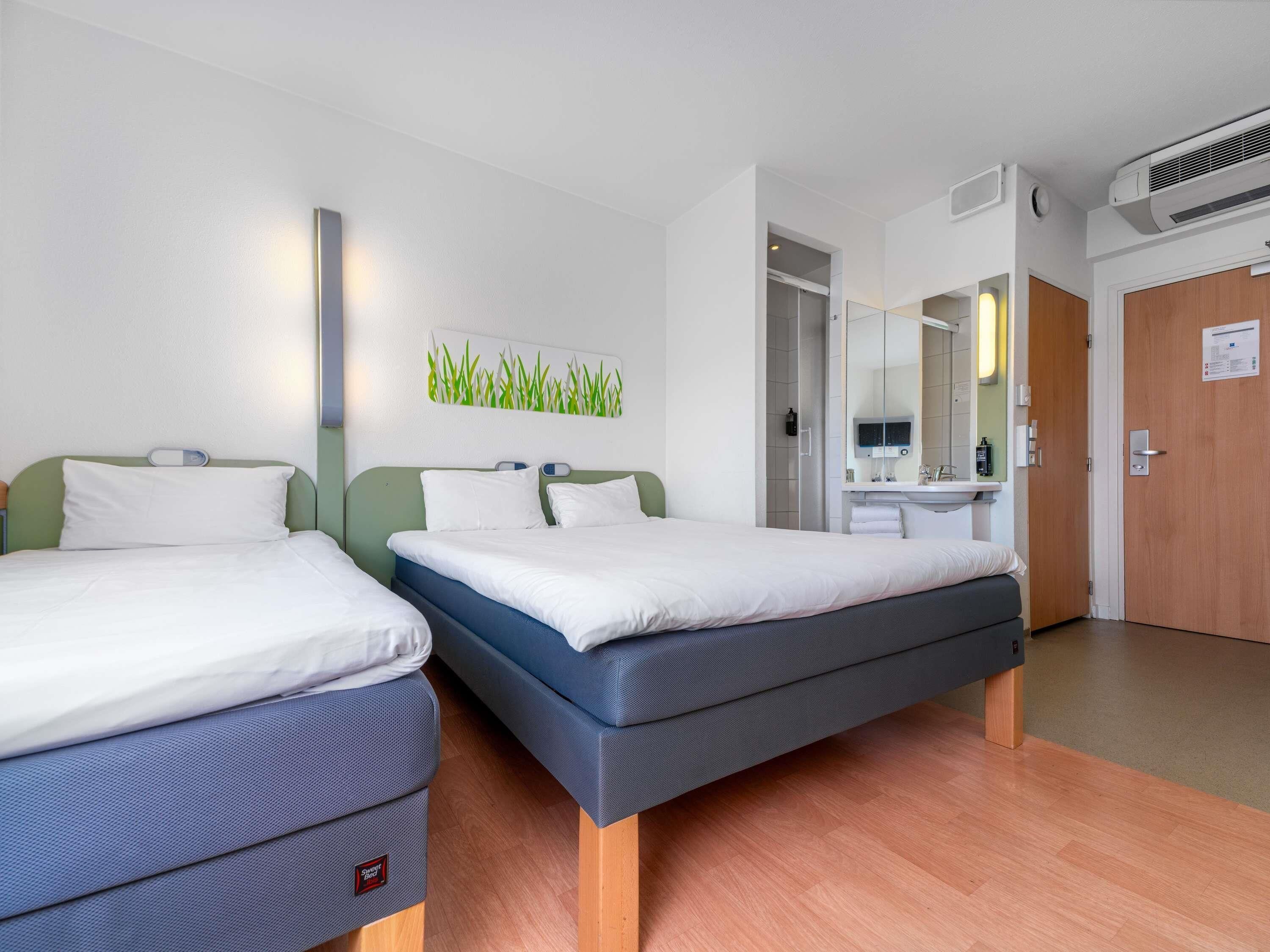 A hotel room with two beds, a bathroom with a mirror, and an air conditioning unit.