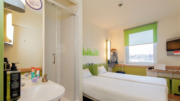 Habitación de hotel con cama, escritorio con computadora portátil, ventana con vistas, baño con lavamanos y espejo, y artículos de tocador en la encimera.