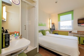 A hotel room with a bed, a desk with a laptop, a window with a view, a bathroom with a sink and mirror, and toiletries on the counter.