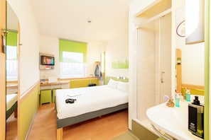 A hotel room with a bed, a desk with a laptop, a TV, a bathroom with a sink and mirror, and a shower.