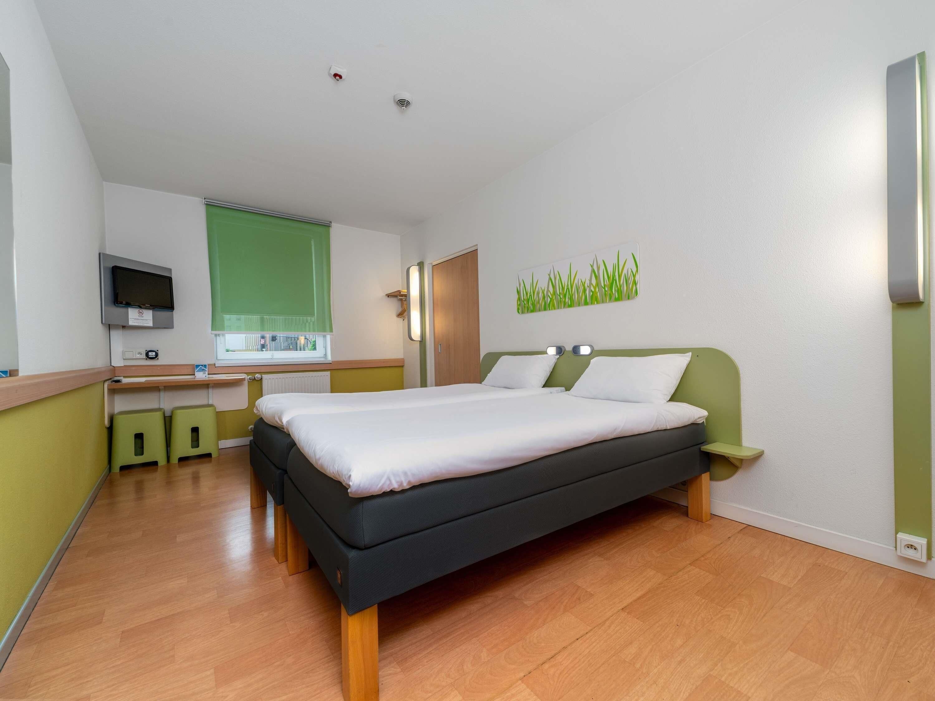 Ein modernes Hotelzimmer mit Bett, Nachttischen, einem Schreibtisch mit Stuhl, einem Fernseher und einem Fenster mit Jalousien.