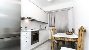 A compact kitchen with white cabinets, a stainless steel refrigerator, an oven, and a dining table with a wooden chair. - Charming 2 bdr apt next to Piraeus port (Piraeus)
