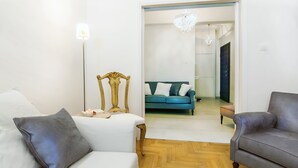 A room with a bed, a chair, a small table with a lamp, and a doorway leading to another room with a couch and a chandelier. - Charming 2 bdr apt next to Piraeus port (Piraeus)