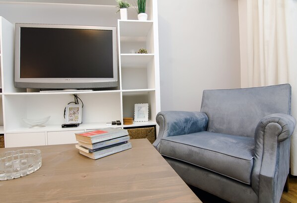 A living room with a flat-screen TV on a white entertainment unit, a grey armchair, and a coffee table with books.