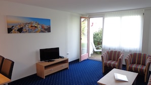 A hotel room with a television on a wooden stand, striped armchairs, a small table, and a painting of a coastal scene. - APART HOLIDAYS Morschach (Morschach)