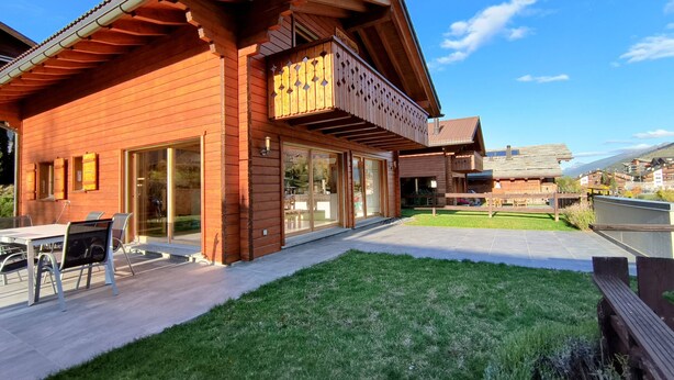 A wooden chalet with large sliding glass doors, a patio with outdoor seating, and a view of the surrounding area.