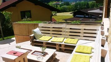 Eine Terrasse mit Holzmöbeln und Kissen, ein kleiner Tisch mit einer Pflanze und Blick auf eine ländliche Landschaft mit Häusern und Hügeln.