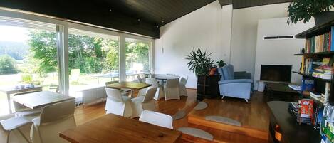 A spacious living area with a wooden dining table, white chairs, a blue sofa, a bookshelf, and a television.