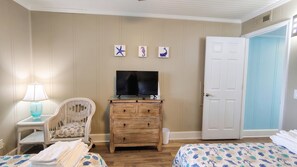 A room with a television on a wooden dresser, a wicker chair, two beds with patterned bedding, and a door.