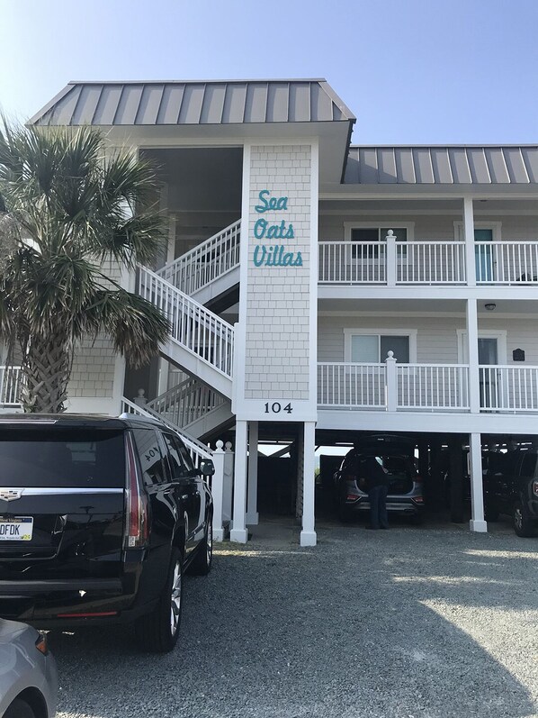A two-story building with a sign reading "Sea Oats Villas" and the number 104. There are multiple parking spaces in front, and a black SUV is parked in one.