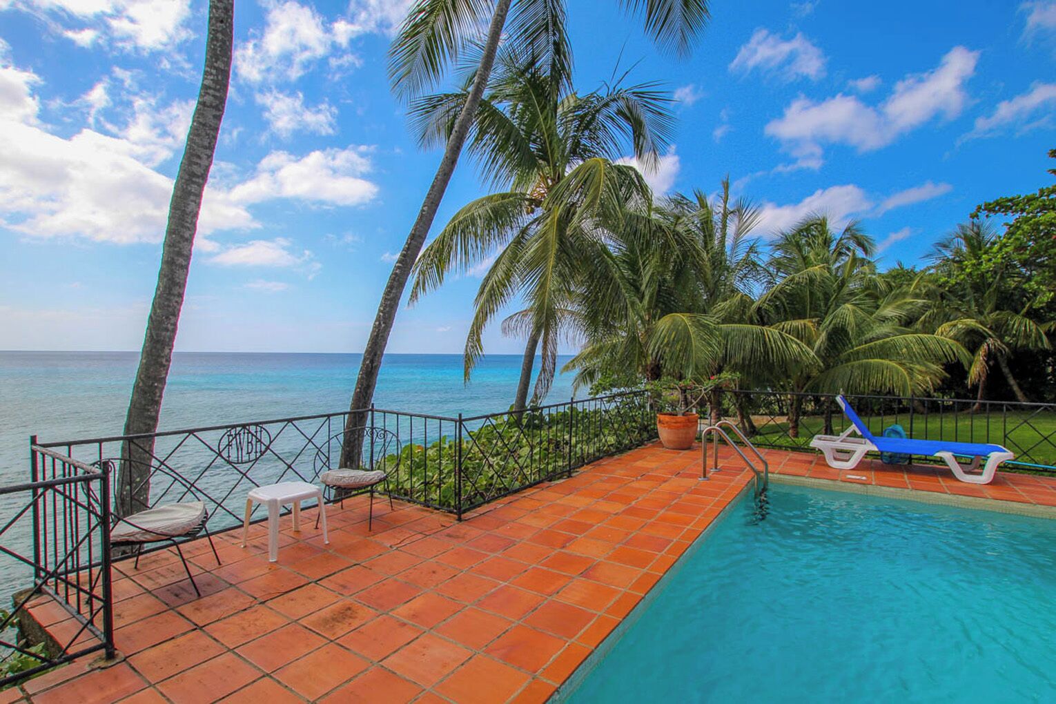 A pool area with a view of the ocean, surrounded by palm trees and a tiled deck.