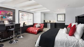 A modern hotel room with a large bed, a desk with a computer, a red leather chair, and a kitchen area.