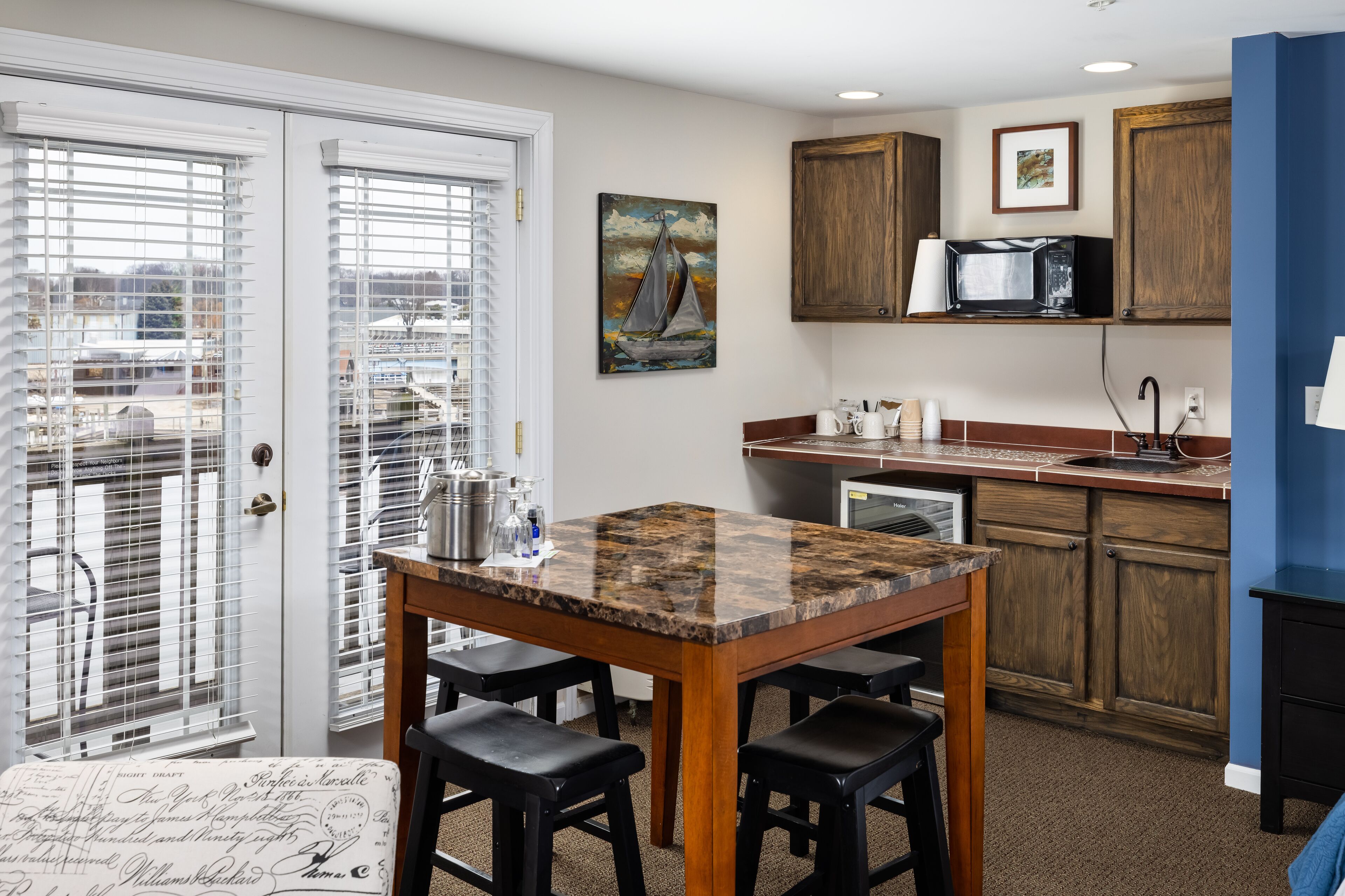 A kitchen with a granite countertop, wooden cabinets, and a microwave. There is a painting of a sailboat on the wall.