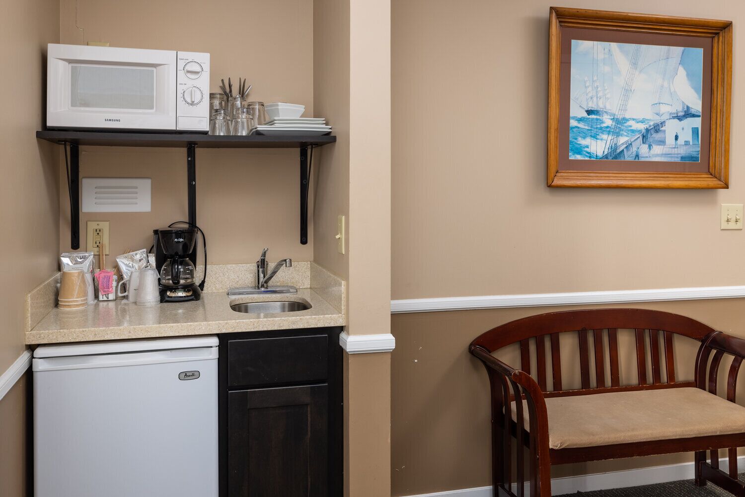 A compact kitchenette with a microwave, coffee maker, and sink. A chair is visible in the foreground.